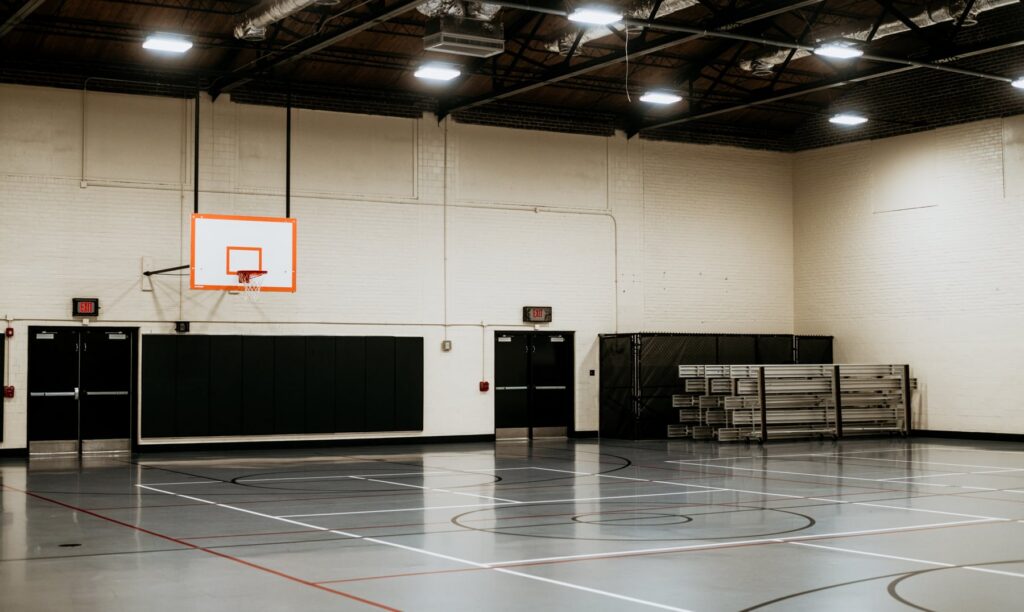 Empty indoor gym with an orange-framed basketball hoop mounted on a white brick wall and stacked bleachers to the right.