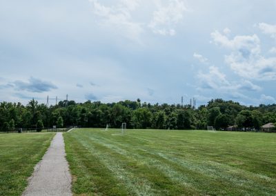 Wide grassy park with a central gravel path, soccer goals along the field, and a tree line under a cloudy blue sky.