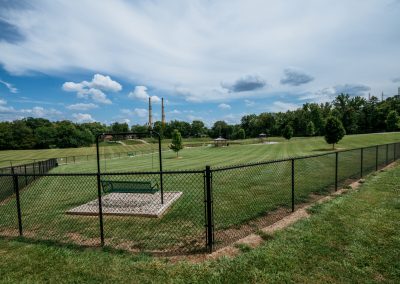 Wide park scene with a black chain-link fence in the foreground, a swing set, bench, and expansive grassy fields; two tall industrial smokestacks rise in the distance beneath a blue sky with scattered clouds.