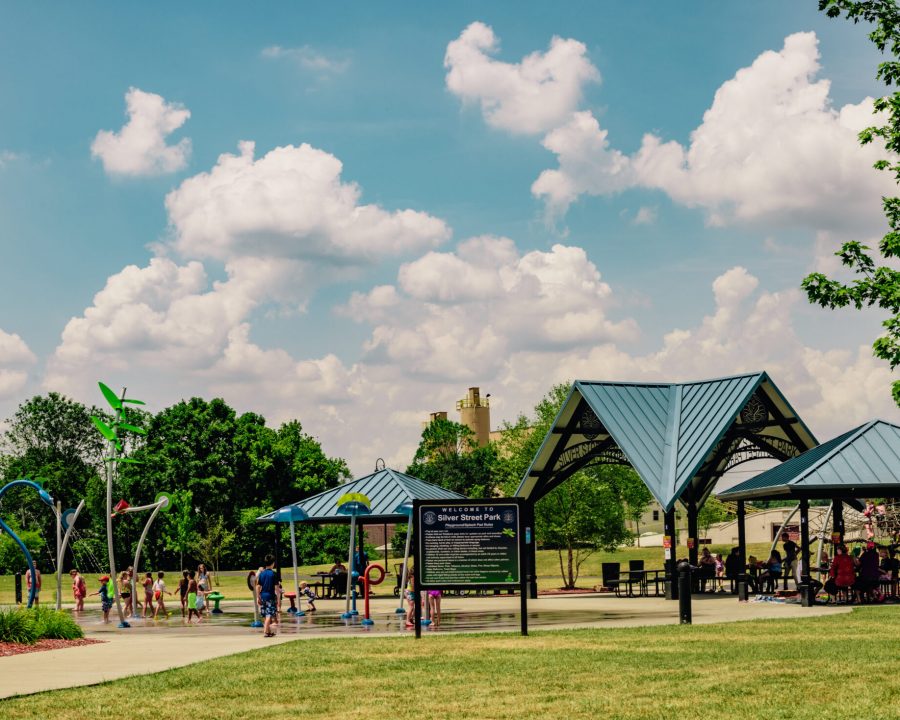 Park scene with children at a splash pad, water jets, and a blue-roof pavilion under a sunny, cloudy sky.