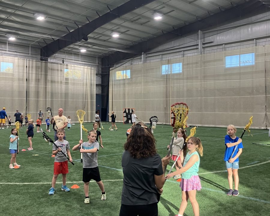 Children practicing lacrosse on an indoor turf field with coaches guiding them; orange cones on the ground and players holding sticks near a coach facing away.